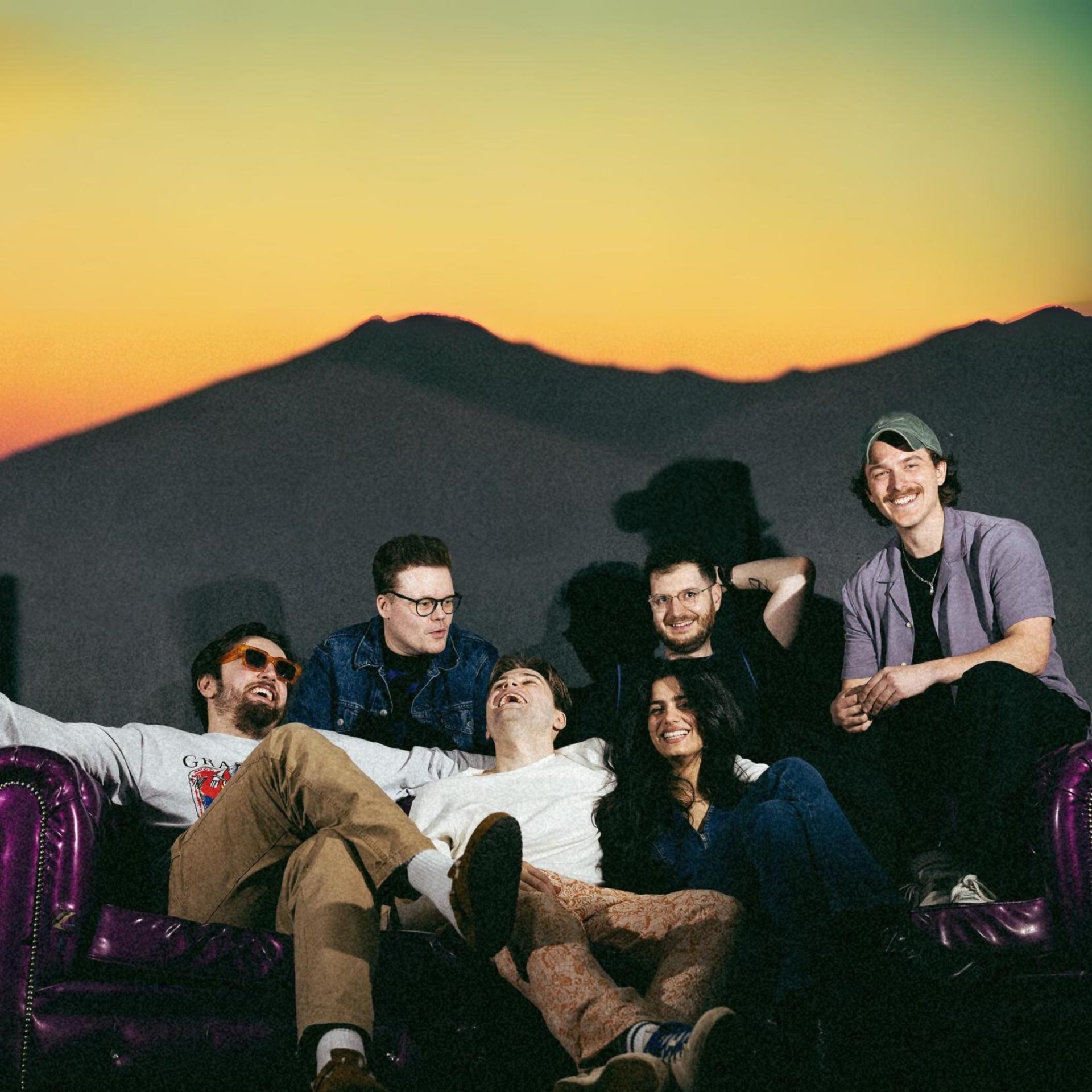 The six members of the band smiling, sitting in front of a mountain backdrop at sunset.