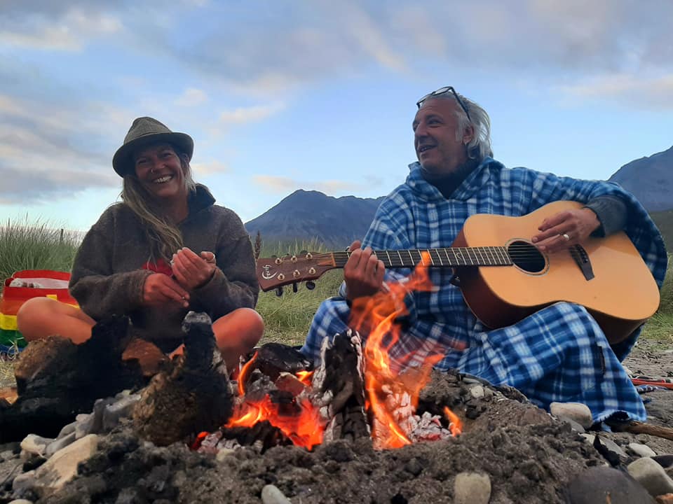 An image of Vik and Ade smiling in front of a campfire with mountains in the background. Ade is holding a guitar.
